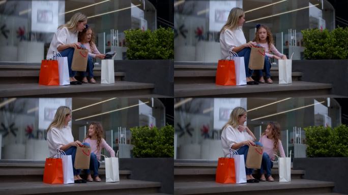 妈妈和女儿坐在商场的楼梯上，看着他们购买的东西，拥抱着，微笑着