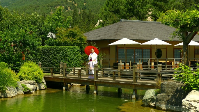 身着和服的年轻女子在茶馆的背景下沿着公园池塘上的木桥行走
