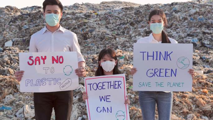 一家三口不同年龄的志愿者站在亚洲垃圾填埋场的巨大垃圾场上，举着为保护自然而战的环保运动积极分子的海报