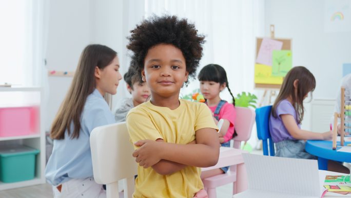 教室里，非洲小男孩快乐地坐在桌子上。可爱的学龄前儿童在幼儿园女教师授课时，交叉手臂看着镜头。
