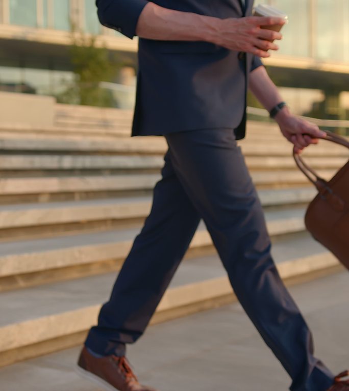 一位身穿蓝色西装、背着皮笔记本电脑包和一杯茶的年轻经理在晚上下班后走下楼梯