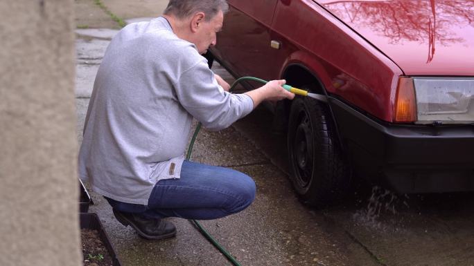 一位老人手里拿着一根软管，在自家后院洗车。