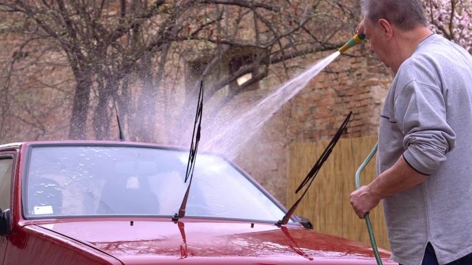 一位老人手里拿着一根软管，在自家后院洗车。
