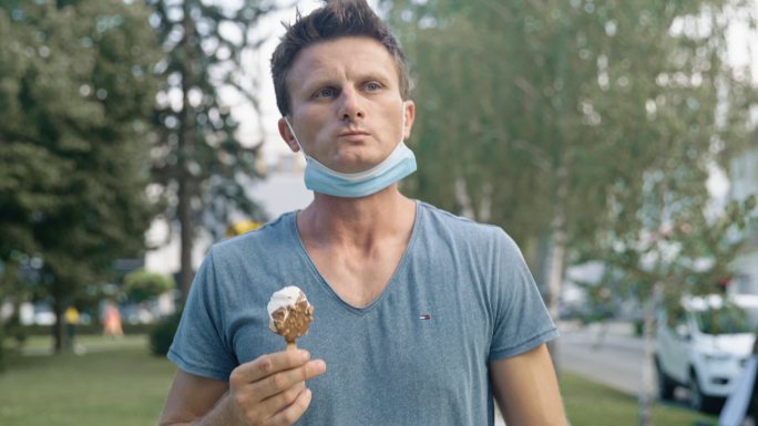 SLO MO男子戴着防护面罩在公园散步时吃巧克力（冰淇淋）冰棍