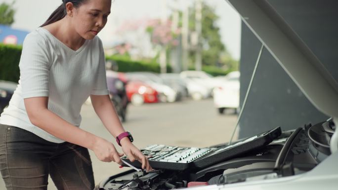 在公寓停车场，一位亚洲中年妇女拿着一个大工具箱，打开、选择并用扳手自己检查汽车电池