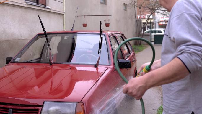 一位老人手里拿着一根软管，在自家后院洗车。