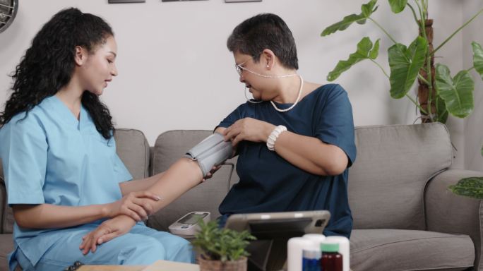 护士在家里的客厅里给一位亚洲老人使用血压监测仪。在家中提供医疗保健服务。护理老年患者。个人护理业务。