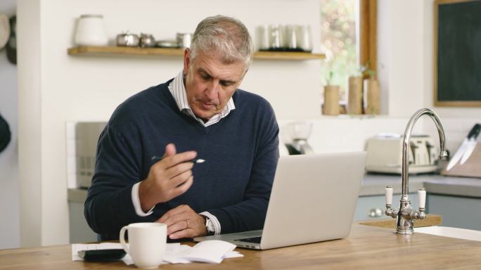 一位老年人在家中使用笔记本电脑，一边计划财务和支付账单。一个成熟的男人正在处理文书工作并使用计算机在