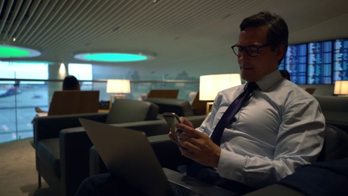 商务人士在VIP机场休息室用笔记本电脑查看智能手机上的电子邮件