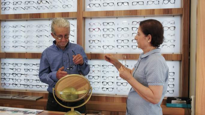 在眼镜店的镜子前，一位年长的女士向她的年长男性顾客解释眼镜