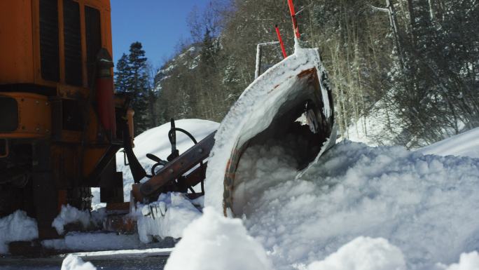 冬日阳光明媚的一天，一辆橙色拖拉机在山间森林旁的雪堆后面犁出了厚厚的雪