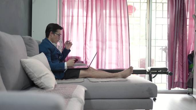 一名亚洲中国男子在家工作，穿着短裤，穿着合适的西装，在网上会议时坐在沙发上使用笔记本电脑。他的孩子们
