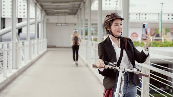 戴着自行车头盔的亚洲美女女商人在穿过中心城区的天桥时停下了自行车，同时愉快地进行视频通话，另一名女子