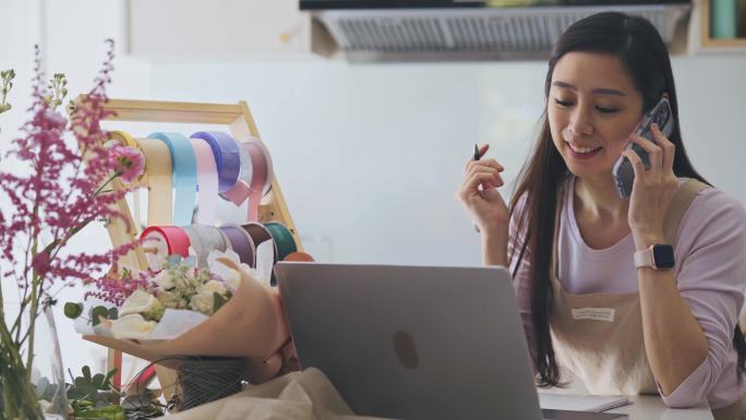亚洲女花艺师，一家小型商业花店的老板，正在用笔记本电脑对付花草。检查库存，接受客户订单，在线销售产品