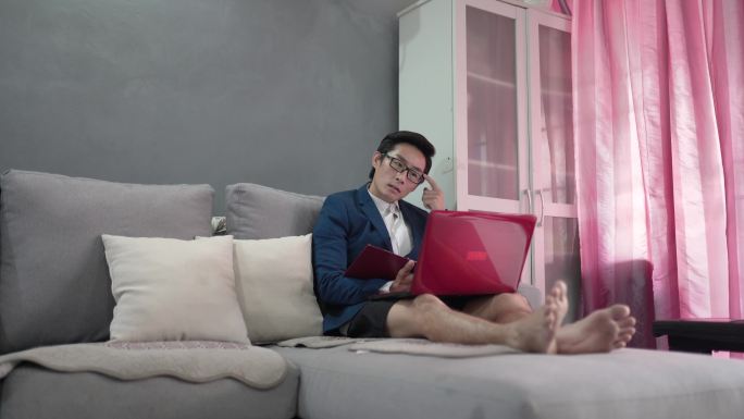 一名亚洲中国男子在家工作，穿着短裤，穿着合适的西装，在网上会议时坐在沙发上使用笔记本电脑。他的孩子们