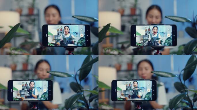 一位年轻的亚洲女性在树店用手机接收订单，并在夜间在线直播植物录制视频。