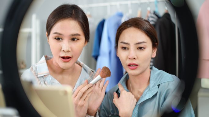 身穿蓝色条纹t恤的亚洲美女手持刷子，在朋友的脸上涂上粉末，展示如何通过社交网络上的直播视频为化妆品销