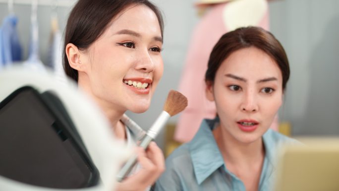 两位可爱的亚洲女博主用相机展示美容产品，并在社交网络上直播视频出售。美容师女孩展示用化妆刷化妆的笑脸