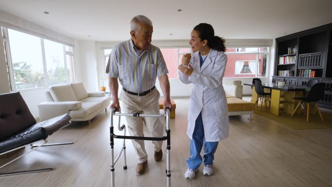 一位拉丁美洲老人在家中，在女医生的帮助下，试图用助行器行走