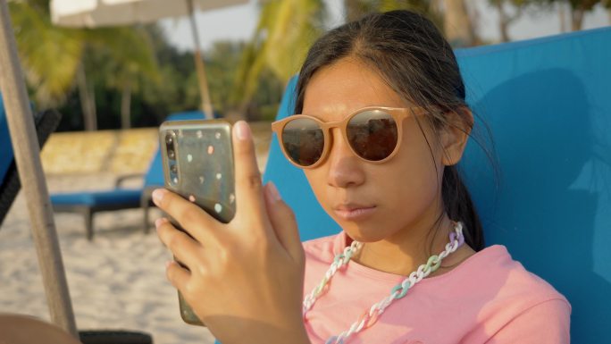 一个亚洲女孩在度假时，早上在沙滩椅上放松时使用智能手机，带着家庭、生活方式的概念。