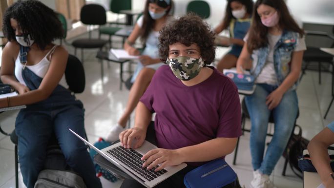 一个十几岁的男孩在教室里用笔记本电脑戴着口罩的照片
