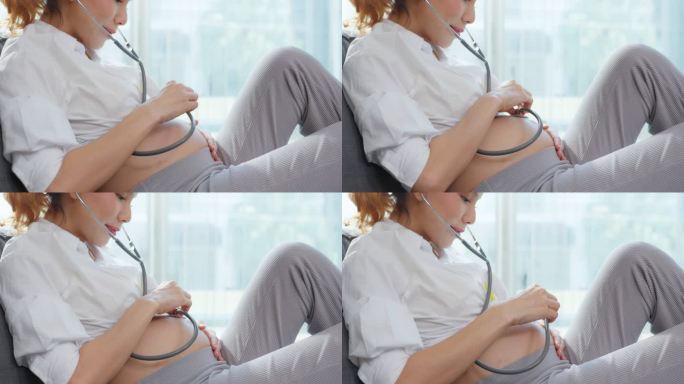 亚洲美女孕妇坐在沙发上，倾听婴儿的心跳。迷人的年轻怀孕母亲在使用听诊器时感到快乐和兴奋，在家里倾听新