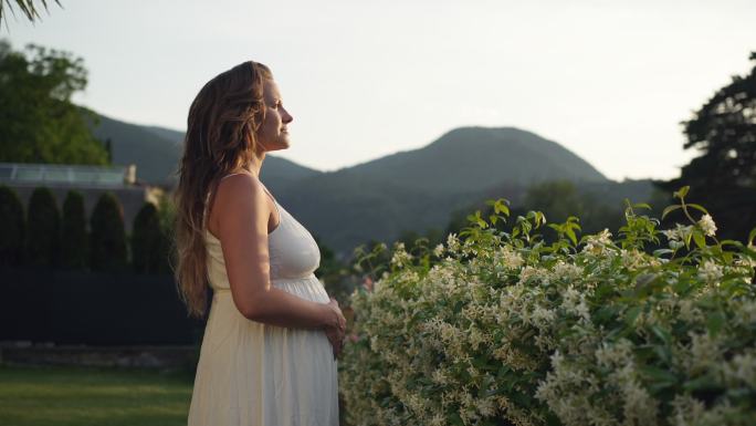 身穿白色连衣裙的孕妇站在花园里的花丛中，抱着她的肚子，看着窗外的景色