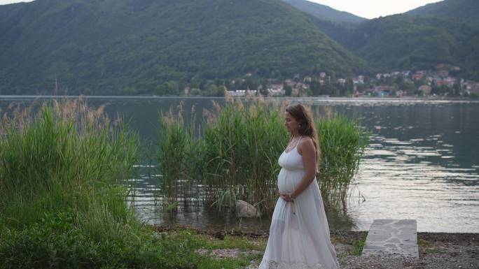 孕妇穿着白色连衣裙，抱着肚子在湖边放松，欣赏群山