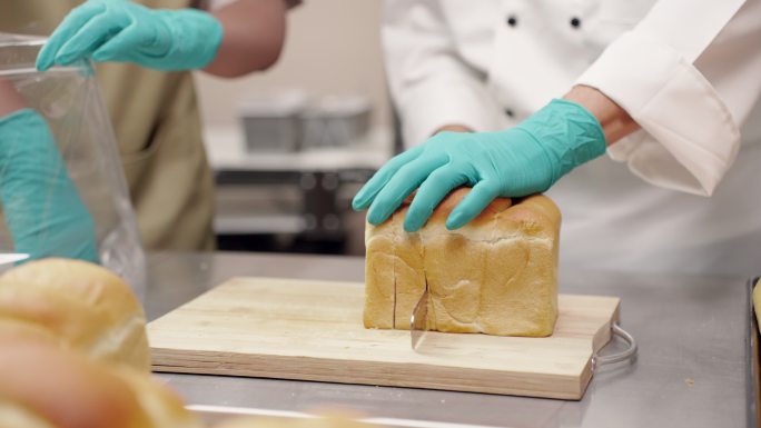 特写：在一家商业厨房，小企业里，厨师戴着手套切面包，助手在将面包送到顾客手中之前帮着将面包装进塑料袋