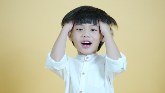 微笑可爱的亚洲男孩表现出兴奋和乐趣，举起手来来回摩擦头发。站在柔和的黄色背景墙上。工作室肖像。人的概