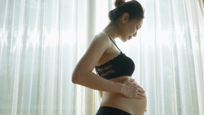 亚洲年轻美丽的孕妇抱着并抚摸她的肚子。迷人的孕妇妈妈站在客厅窗户附近的室内，怀着幸福和爱看着肚子