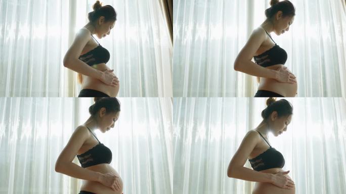亚洲年轻美丽的孕妇抱着并抚摸她的肚子。迷人的孕妇妈妈站在客厅窗户附近的室内，怀着幸福和爱看着肚子