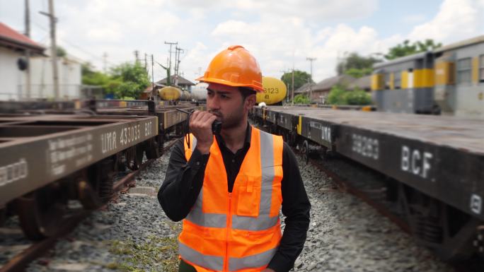 亚洲工程人员戴着头盔和制服在火车站上检查火车。男工程专家使用步行对讲机检查货运列车。