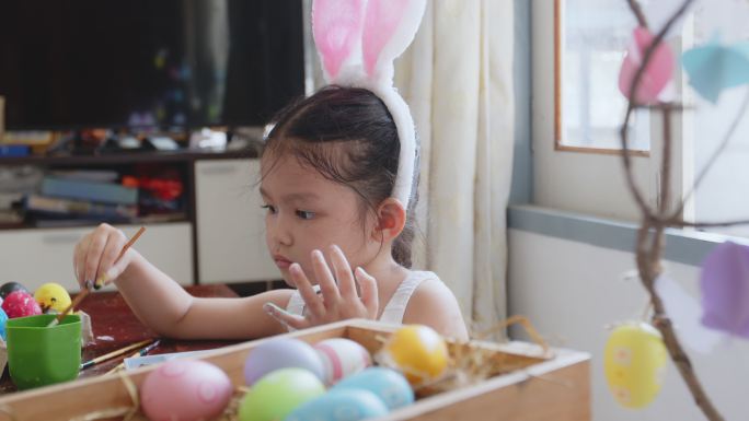 可爱的亚洲女孩戴着兔子头带，开心地给她的手上色。孩子们玩得很开心，为复活节做准备，画鸡蛋和装饰鸡蛋。