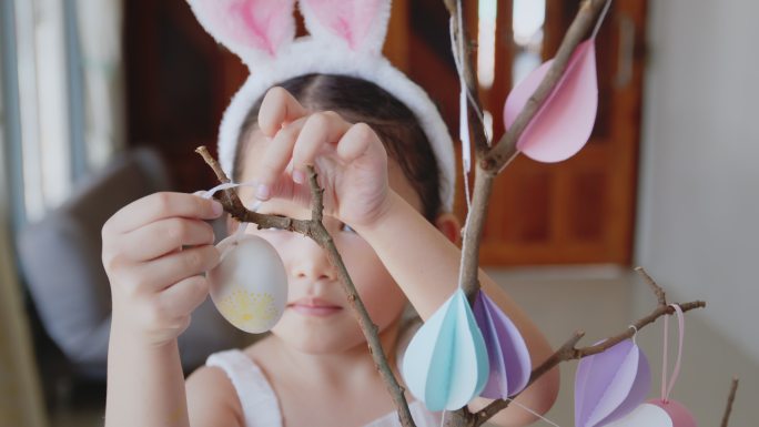一个亚洲小女孩把五颜六色的复活节彩蛋挂在树枝上，帮父母装饰复活节的房间。