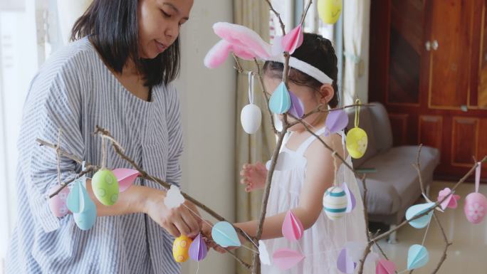 一个亚洲小女孩把五颜六色的复活节彩蛋挂在树枝上，帮父母装饰复活节的房间。