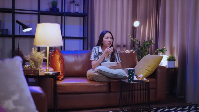 亚洲女性单身女性晚上坐在家里客厅的沙发上，端着一碗爆米花零食看网络电视