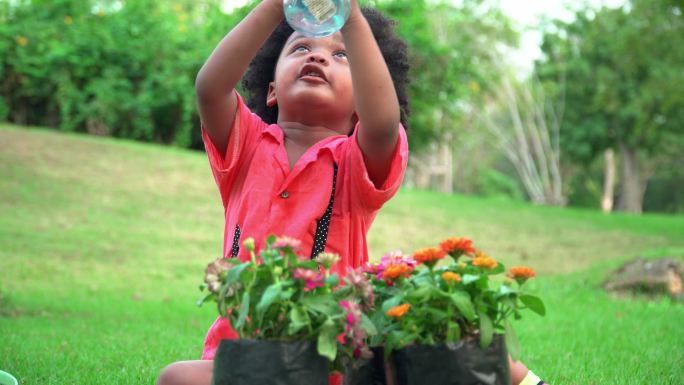 美国非洲小男孩，留着非洲式发型，坐在绿色公园的草地上，用喷雾瓶将水喷洒成气雾剂。孩子在花园里玩水，花