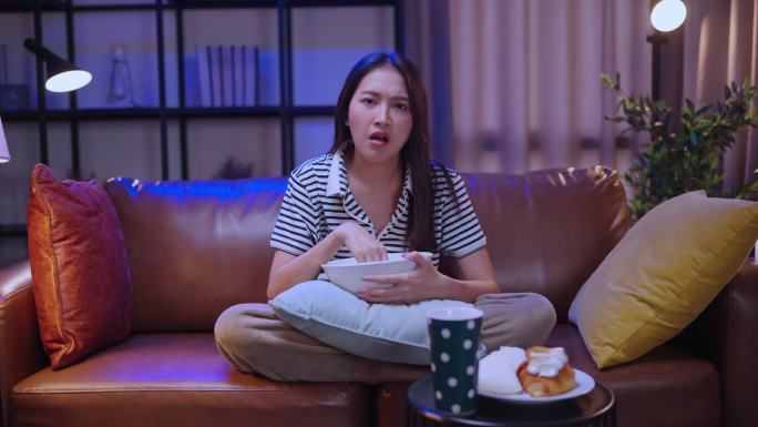 亚洲女性单身女性晚上坐在家里客厅的沙发上，端着一碗爆米花零食看网络电视