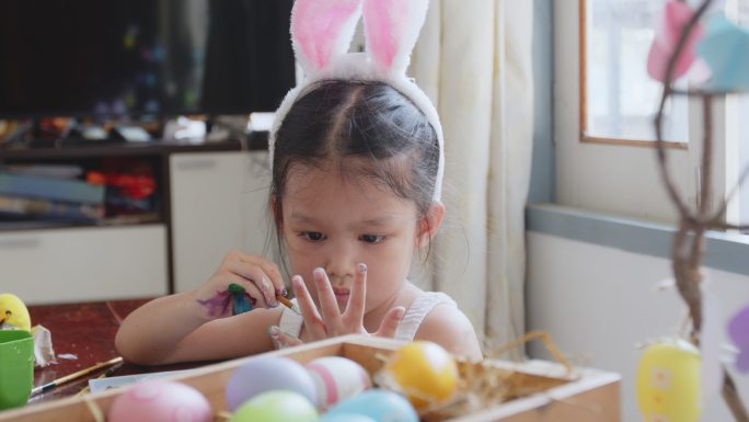 可爱的亚洲女孩戴着兔子头带，开心地给她的手上色。孩子们玩得很开心，为复活节做准备，画鸡蛋和装饰鸡蛋。
