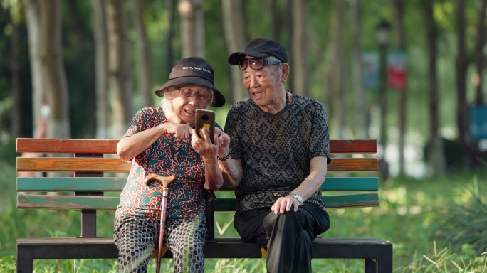 幸福的老人夫妇坐在公园长椅一起看手机聊天