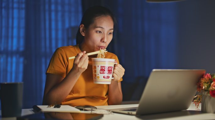 一名亚洲妇女晚上在家里用笔记本电脑工作时吃方便面
