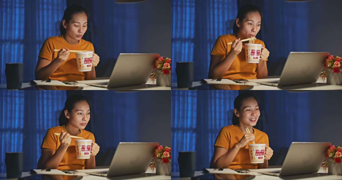 一名亚洲妇女晚上在家里用笔记本电脑工作时吃方便面