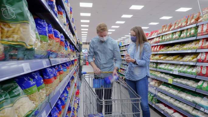 戴着口罩的年轻夫妇用购物车在超市过道里购买食品杂货