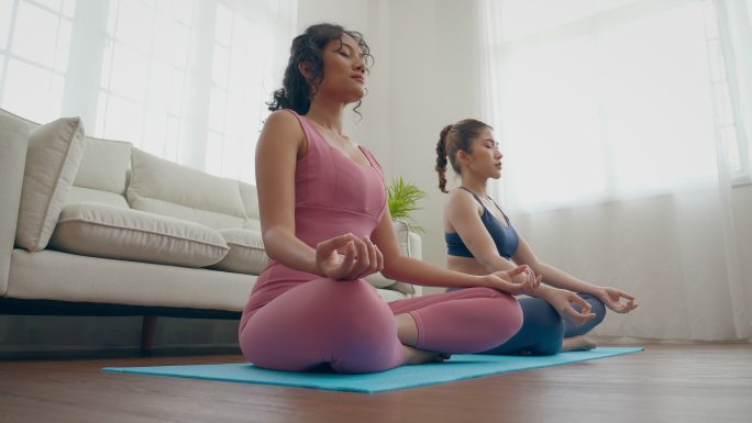 一对年轻的亚洲女性夫妇穿着胸罩和紧身裤，坐在家里的垫子上练习瑜伽和冥想