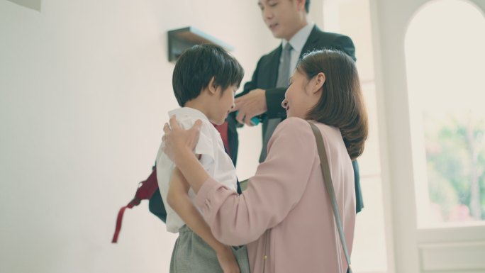 亚洲母亲在准备送小儿子上学之前拥抱着小儿子，而亚洲父亲则在家里的客厅里背着儿子准备上学的背包。亲子关