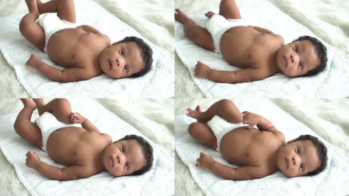 高角度视角：非洲男婴的微笑，天真的婴儿儿子，娇生惯养的躺在卧室的白色床上，带着怀疑的心情看着镜头。父