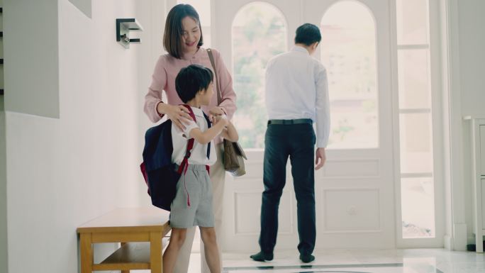 一位亚裔父亲早上上学前给儿子系鞋带。可爱的小男孩背着双肩包，得到妈妈的帮助。幸福的家庭，亚裔家长和他