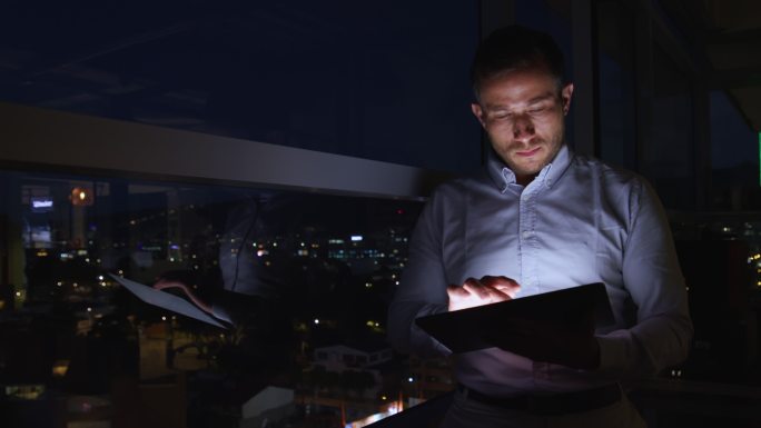 一位拉丁美洲商人在办公室工作到很晚，他使用平板电脑，看着窗外的景色，非常体贴