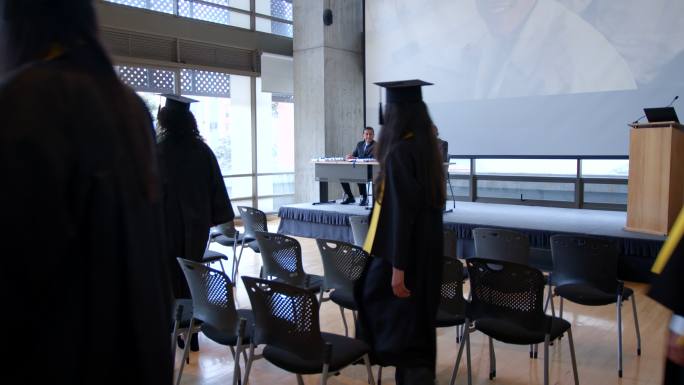 一群学生走进他们的毕业典礼，而董事会成员坐在桌子旁边，手里拿着所有的毕业证书